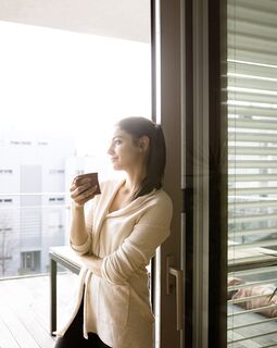Junge Frau mit Kaffeetasse lehnt an geöffneter UNILUX Holz-Alu Hebe-Schiebe Anlage mit Blick auf Balkon