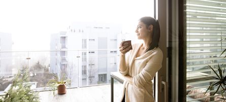 Junge Frau mit Kaffeetasse lehnt an geöffneter UNILUX Holz-Alu Hebe-Schiebe Anlage mit Blick auf Balkon