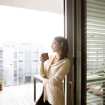 Junge Frau mit Kaffeetasse lehnt an geöffneter UNILUX Holz-Alu Hebe-Schiebe Anlage mit Blick auf Balkon